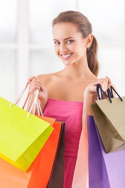 Good day for shopping. Beautiful young woman in pink dress holding shopping bags and smiling at camera