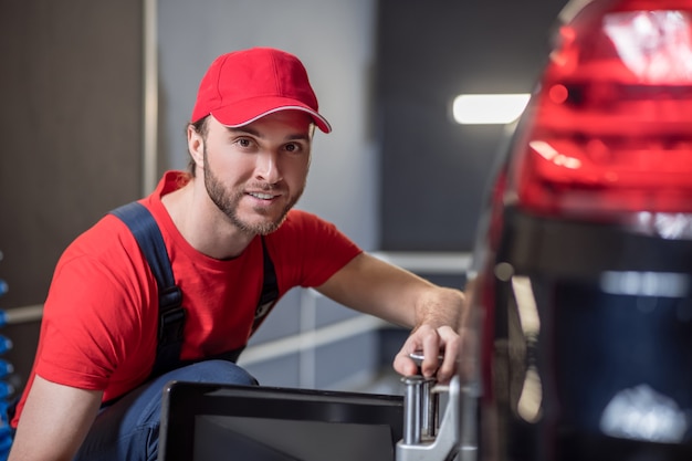 Good day, work. Happy young bearded man in red cap and tshirt working crouched near car wheel in workshop