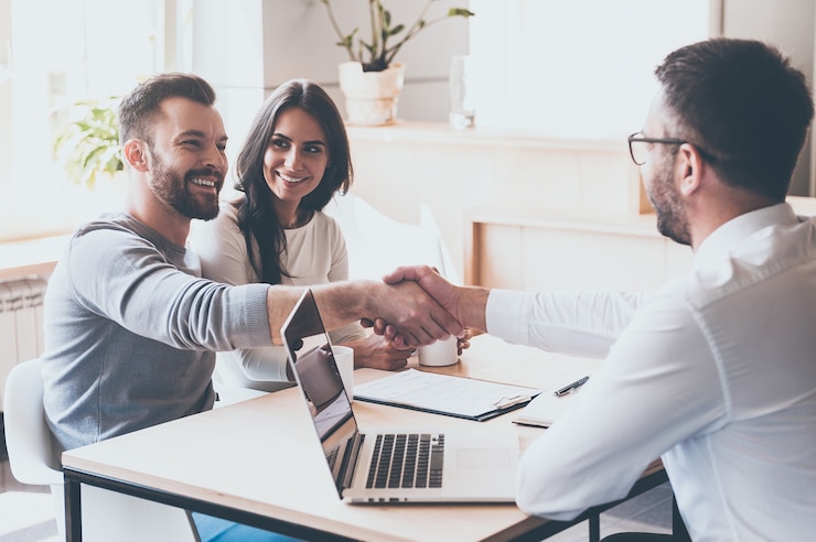 Photo good deal! cheerful young man bonding to his wife while shaking hand to man sitting in front of him at the desk