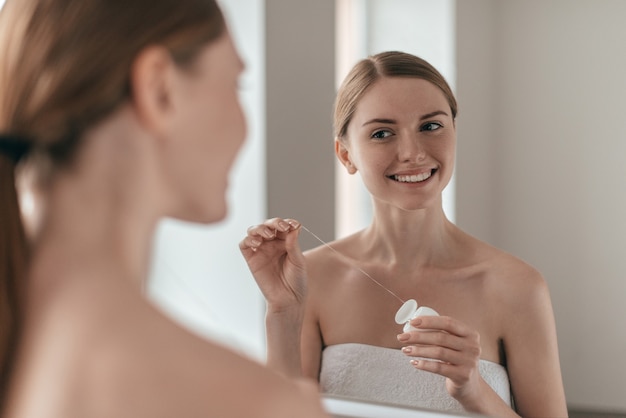 Good habit. over the shoulder view of young beautiful woman\
holding dental floss and smiling while looking in the mirror