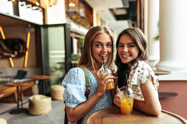 Giovani donne bionde e brune di buon umore in camicette floreali alla moda sorridono sinceramente, bevono limonata e riposano in un ristorante di strada