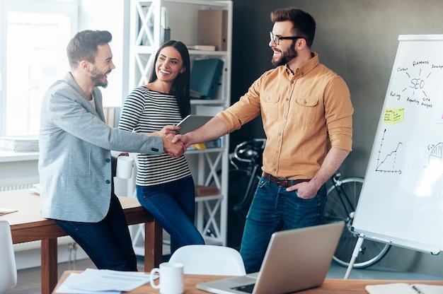 Good job! Confident young man standing near whiteboard and shaking hand to his colleague while young woman standing near them and smiling