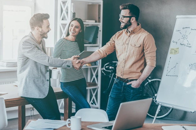 Good job! Confident young man standing near whiteboard and shaking hand to his colleague while young woman standing near them and smiling