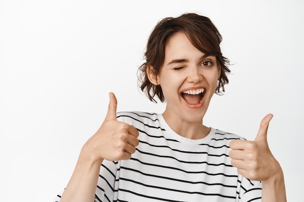 Good job, excellent choice. Smiling brunette girl showing thumbs up and winking, look pleased, standing over white background