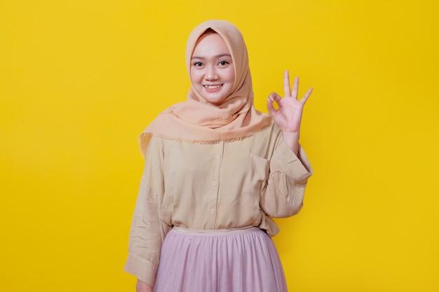 Фото Хорошая работа, гордая счастливая азиатская молодая женщина в хиджабе показывает нормальный жест, подмигивая радостным, изолированным на желтом фоне