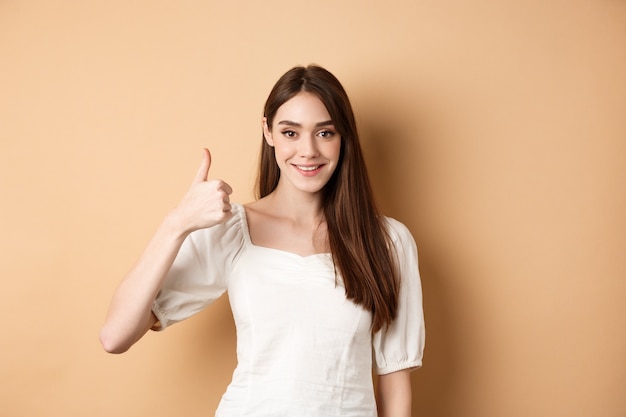 Good job. Smiling candid woman in blouse showing thumb up, praise nice choice, recommending product, standing satisfied on beige background.