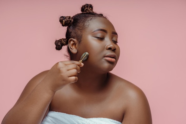 Good looking african woman with closed eyes doing face roller massage isolated on pink background Beauty procedures concept