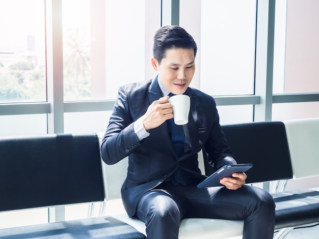 Good looking Asian businessman in suit watching tablet on his hand and drinking white cup of hot coffee while sitting on waiting chair