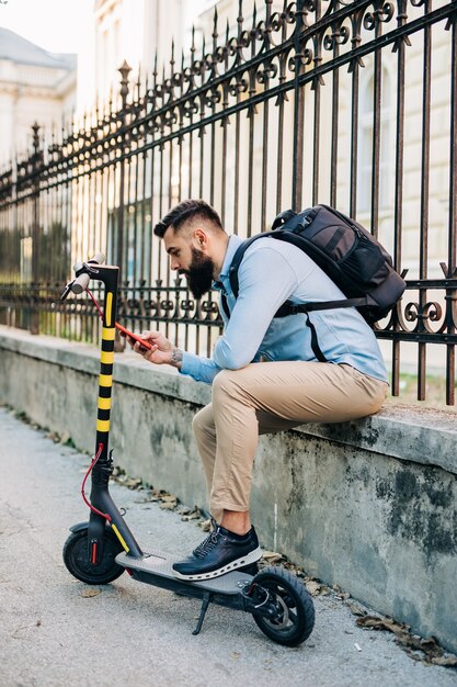 電動スクーターの隣に座って携帯電話を見ている格好良いひげを生やした男。