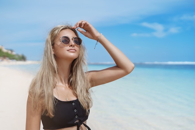 Красивая блондинка загорелая женщина позирует на песчаном пляже возле синего океана