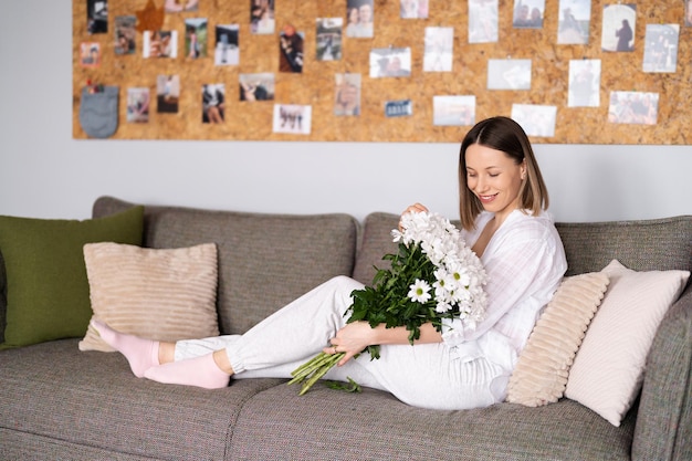 Photo good looking caucasian woman enjoy flowers she is happy to get a fresh bouquet of white chrysanthemum while relaxing on the sofa