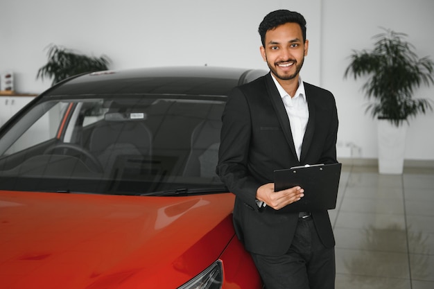 Good looking cheerful and friendly indian salesman poses in a car salon or showroom