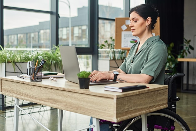 체어에 앉은 장애를 가진 잘 생겼고 자신감 있는 여성이 사무실에서 노트북에서 열심히 일하고 있습니다.
