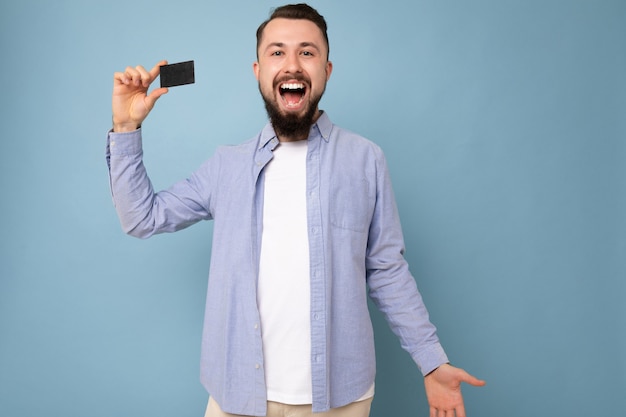 Good looking cool overjoyed smiling brunette bearded young man wearing stylish blue shirt and white t-shirt isolated over blue background wall holding credit card looking at camera and having fun.