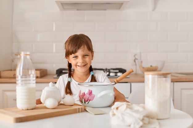 Bella bambina con trecce che indossa abiti in stile casual seduta al tavolo in una cucina leggera e prepara torte o biscotti da sola, circondata da prodotti.