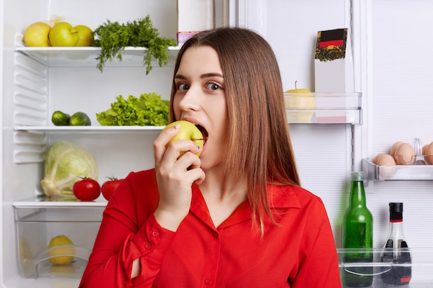 Good looking female model bited delicious apple, stands near opened fridge full of healthy products, demonstares her good nutrition