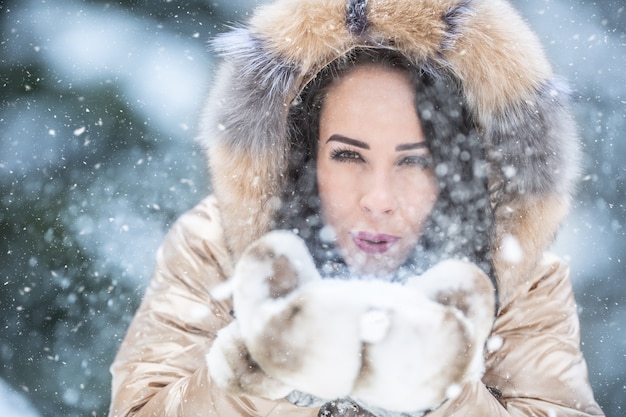 Foto una bella ragazza soffia la neve verso la telecamera vestita con vestiti caldi e pelosi per il freddo inverno.