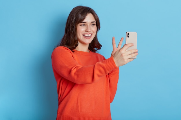 Good looking glad female with toothy smile, makes peace sign or V gesture at camera of cell phone