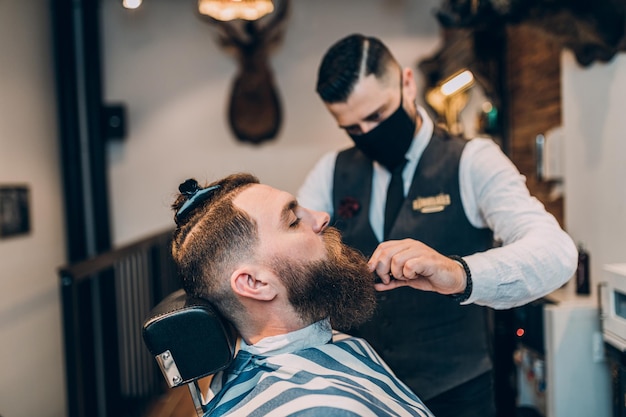 Giovane uomo barbuto bello hipster che visita parrucchiere nel negozio di barbiere. il barbiere indossa una maschera protettiva per il viso a causa del coronavirus.