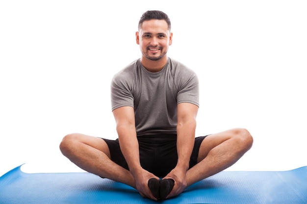 Good looking Latin guy sitting on a yoga mat and doing some stretching exercises on a white background