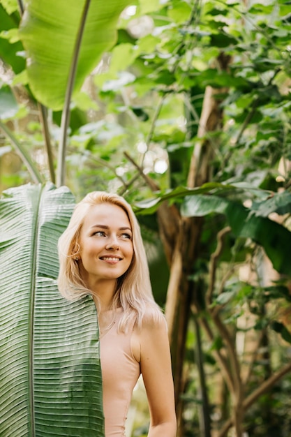 멋진 살색 수영복을 입은 멋진 금발 여성은 자연을 즐기고 숲에서 포즈를 취하고 이국적인 식물의 큰 잎 뒤에 날씬한 몸을 덮고 있습니다. 패션 모델, 뷰티 컨셉입니다.