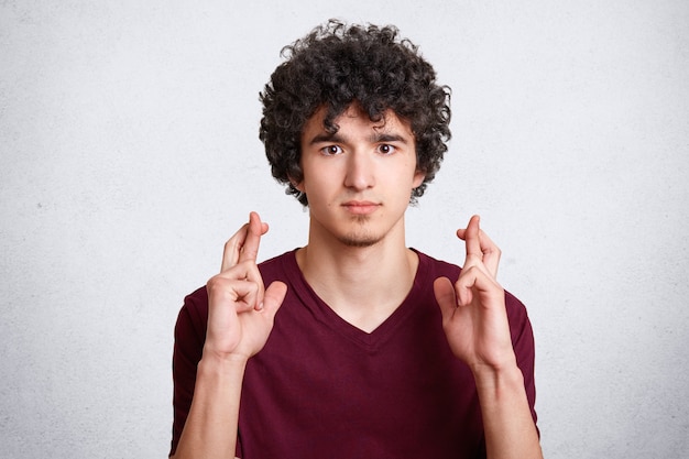 Uomo di bell'aspetto con i capelli croccanti, incrocia le dita, indossa una maglietta casual, ha un'espressione desiderabile, posa contro il muro di cemento bianco