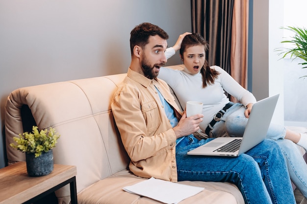 Good looking man in orange shirt and blue jeans and attractive young woman in white turtleneck together saw something shocking on laptop screen