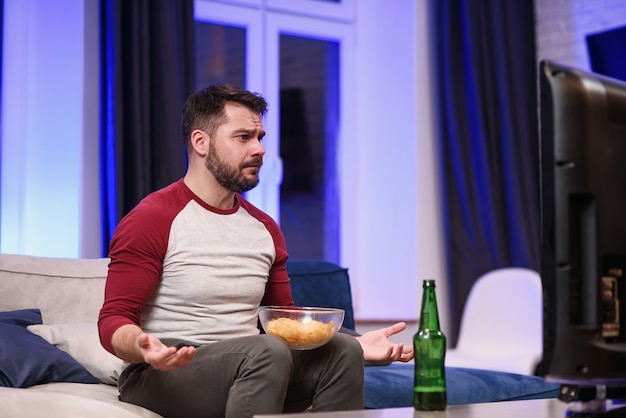 Good-looking modern positive guy with well-groomed beard savoring chipps during emotional review of favourite football team's game on tv at home