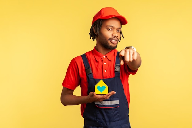 Good looking mover or handyman in red cap and blue overalls holding paper house in hands and pointing finger to camera offering home repair services Indoor studio shot isolated on yellow background