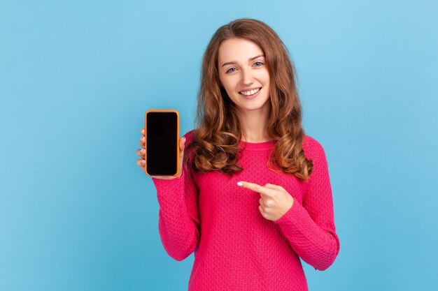 Photo good looking positive happy woman wearing pink pullover showing phone with empty screen pointing cell phone with space foe promotion indoor studio shot isolated on blue background