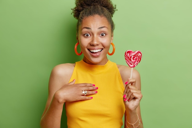 Good looking positive young woman with curly hair keeps hand on
chest smiles happily holds heart shaped lollipop wears yellow
summer t shirt isolated over green background sweet tooth
concept