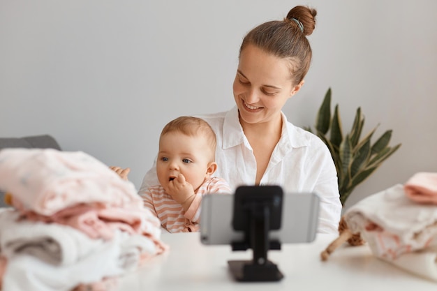 Good looking pretty young female blogger, with hair bun broadcasting livestream at home together with her infant daughter, looking at cute kid, smiling, filming content for her blog.