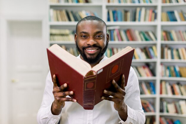 Giovane ragazzo barbuto africano soddisfatto bello in camicia casuale bianca, posante sullo spazio degli scaffali di libro in biblioteca moderna