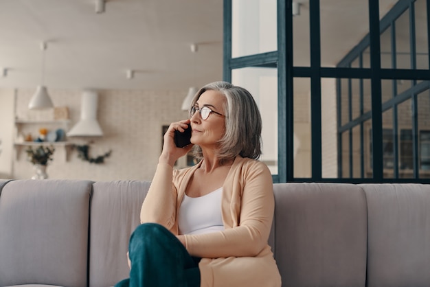 Bella donna anziana in abbigliamento casual che parla allo smartphone mentre è seduta sul divano di casa