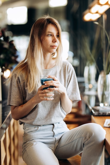 A good looking slim blonde girl,wearing casual style, sits on the chair and holds a cup of coffee in a cozy coffee shop. .