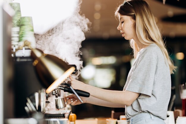 カジュアルな服装の長い髪の格好良いスリムなブロンドは、モダンなコーヒーショップでコーヒーを調理しています。コーヒーを作るプロセスが示されています。 。