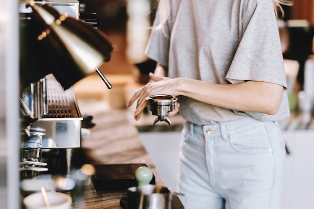 カジュアルな服装の長い髪の格好良いスリムなブロンドは、モダンなコーヒーショップでコーヒーを調理しています。コーヒーを作るプロセスが示されています。 。