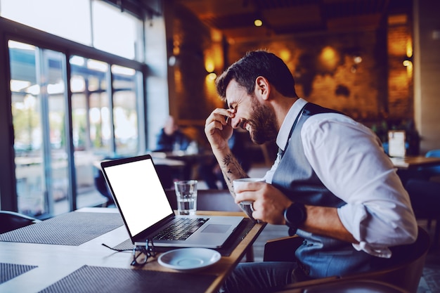 Good-looking smiling caucasian bearded businessman in suit sitting in cafe, holding cup with coffee and laughing. On table are glasses, laptop and water.