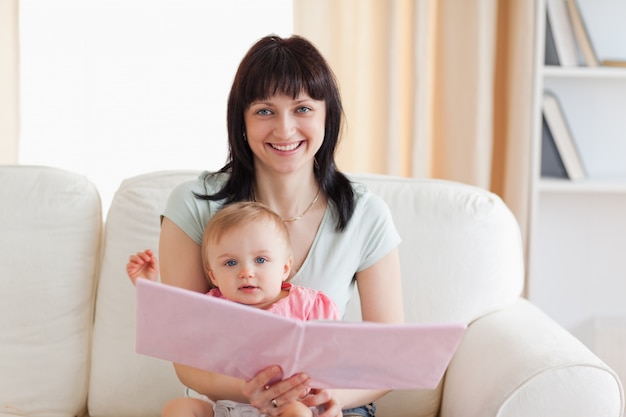 Good looking woman holding her baby and a book in her arms while sitting on a sofa