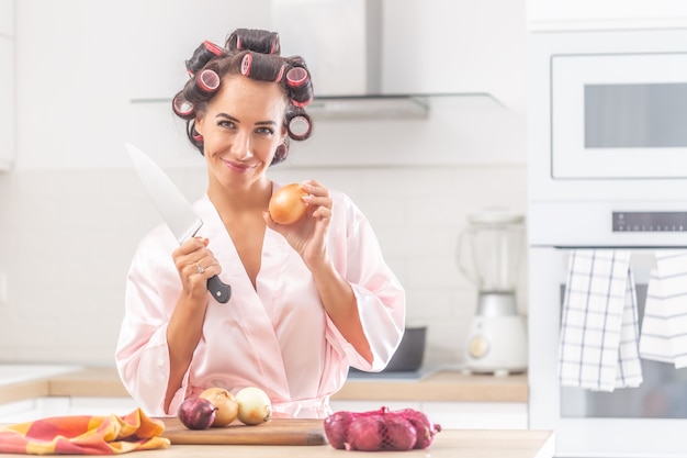 Good looking woman stands in the kitchen holding knife and an onion, wearing hair curlers and night gown.