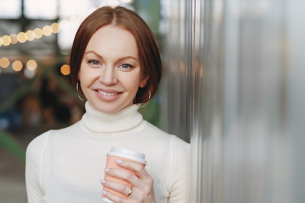 캐주얼한 흰색 터틀넥 스웨터를 입은 잘 생긴 젊은 여성이 향기로운 카푸치노 종이컵을 들고 있거나 커피는 여유 시간이 있는 외부 카메라 포즈를 행복하게 보입니다.