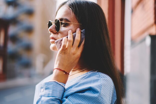 Good looking young female in sunglasses talking on smartphone standing on street