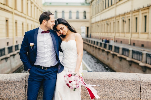 Good looking young female with appealing appearance wears white\
wedding dress holds nice bouquet leans at lovers shoulder enjoy\
togetherness and calm atmosphere as stand on bridge alone