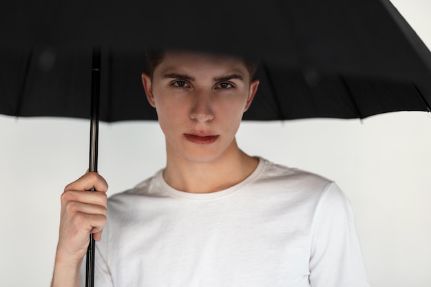 Good-looking young man in stylish casual t-shirt with fashion
black umbrella isolated on white background in studio. pretty
handsome guy takes cover by an umbrella in room. youth summer
menswear.