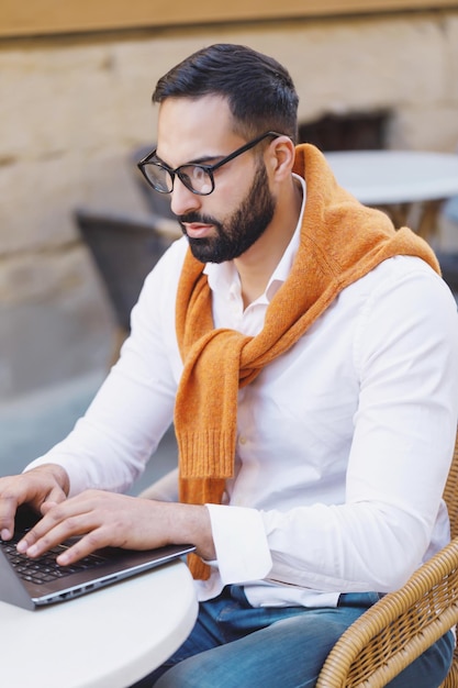 Симпатичный молодой человек, использующий компьютер и держащий руку на подбородке, сидя в кафе на открытом воздухе