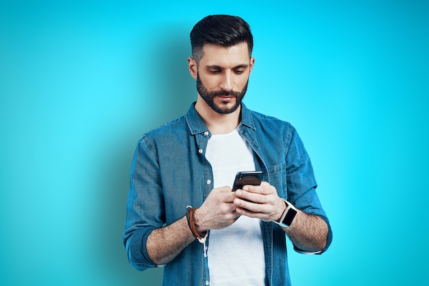 Good looking young man using smart phone while standing against blue wall