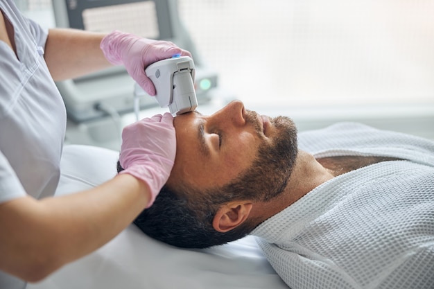 Good-looking young man with stubble lying on daybed while receiving laser facial treatment at wellness center