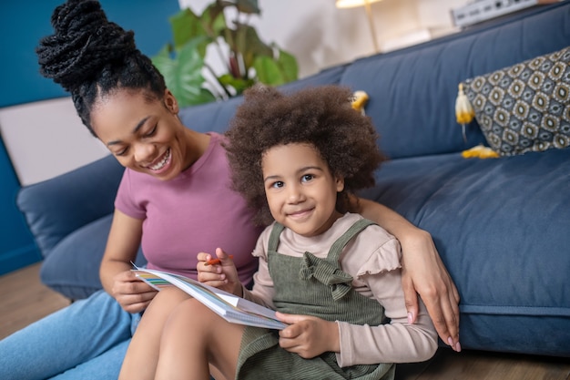 Good moments. Joyful dark-skinned young mother with her little cute daughter drawing with color pencils sitting on floor at home