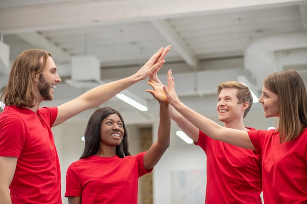 Good moments. Two young girls and guys in identical tshirts touching palms of raised hands standing indoors in an enthusiastic mood