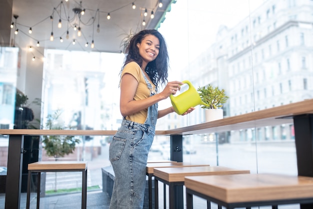 Good mood. Cheerful young long-haired mulatto woman watering flowerpot on table in cafe in afternoon